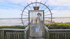 A wooden boardwalk leading to a metal gate with a palm tree design, overlooking a beach. - Seascape 2317 Studio Bedroom Condo by RedAwning (Galveston)
