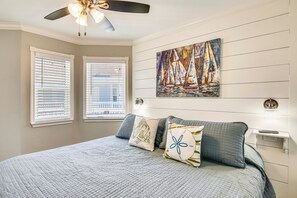 Une chambre à coucher avec un lit, deux fenêtres, un ventilateur de plafond et un tableau représentant des voiliers.