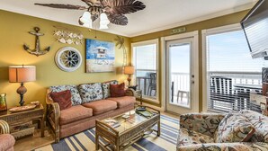 A living room with a sofa, coffee table, and decorative items. There is a ceiling fan, a painting, and a sign that says "BEACH DAY". - Seascape 1209 2 Bedroom Condo by RedAwning (Galveston)