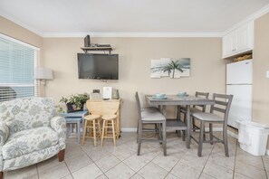 Un espace de vie compact comprenant un coin repas, un téléviseur à écran plat fixé au mur, une petite table avec des chaises, un canapé et un réfrigérateur.