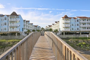 A wooden boardwalk leading to a row of multi-story buildings with balconies, likely part of a hotel or resort. - Seascape 2105 1 Bedroom Condo by RedAwning (Galveston)