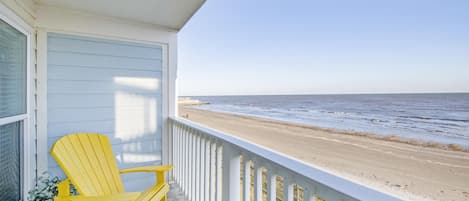 A balcony with a view of the beach, featuring two yellow Adirondack chairs and a white railing.