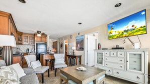 A modern kitchen with wooden cabinets, a dining area with a wooden table and chairs, and a living room with a sofa and a television mounted on the wall.