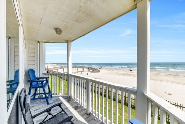 A covered porch with a view of the beach, featuring a wooden deck and blue chairs.