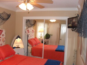 A bedroom with two beds, a ceiling fan, a TV, and a mirror. - Seascape 2318 1 Bedroom Apts by RedAwning (Galveston)