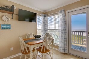 A dining area with a table and chairs, a television mounted on the wall, and a view through the window. - Seascape 2318 1 Bedroom Apts by RedAwning (Galveston)