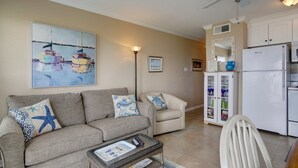 Een woonkamer met een beige bank, een salontafel, een witte fauteuil, en een schilderij van boten aan de muur.