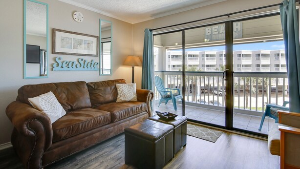 Ett vardagsrum med en brun lädersoffa, en liten svart pall och en blå fåtölj. Ett stort fönster med utsikt över en byggnad med texten ”BEACH” syns.