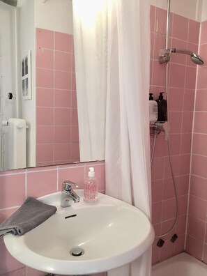 Une salle de bain avec un lavabo blanc, un miroir, une douche équipée d’un pommeau de douche à main et des murs recouverts de carrelage rose.