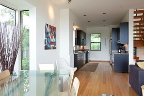 Una cocina moderna con gabinetes oscuros, un comedor con mesa de vidrio y una escalera con barandilla de madera.