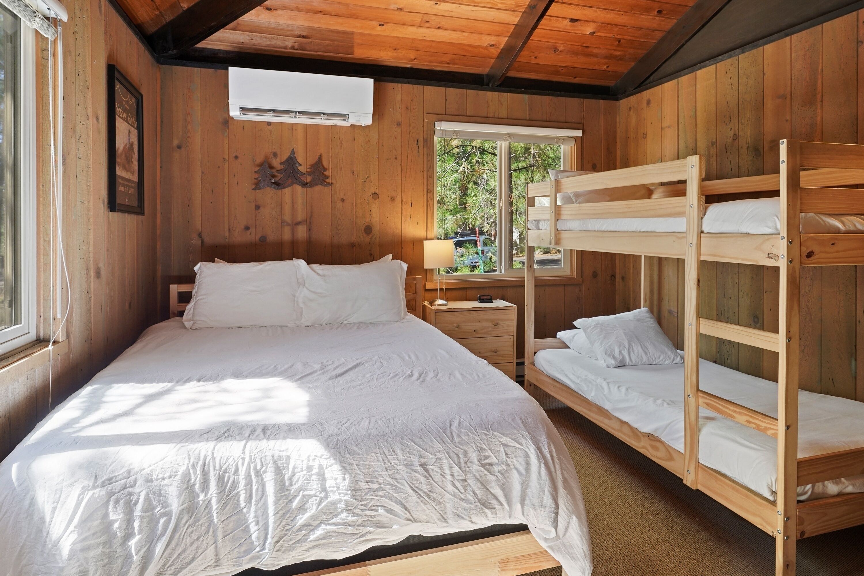 A wooden cabin room with a bunk bed, a single bed, a nightstand, and a window with a view of trees.