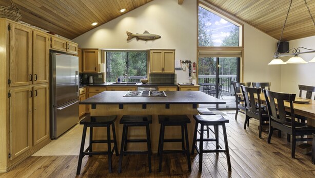 A spacious kitchen with wooden cabinets, a large island with bar stools, a fish hanging on the wall, and large windows offering views of the outdoors. - 2 Quail Home Private Hot Tub, Steps from SHARC and short walk to the Sunriver Village by RedAwning (Bend)