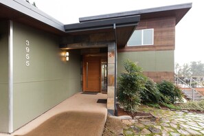 Ein modernes Haus mit einem Kiesweg, der zu einer Holztür führt, mit der Hausnummer 3955 an der Wand und einem kleinen Gartenbereich mit Sträuchern.