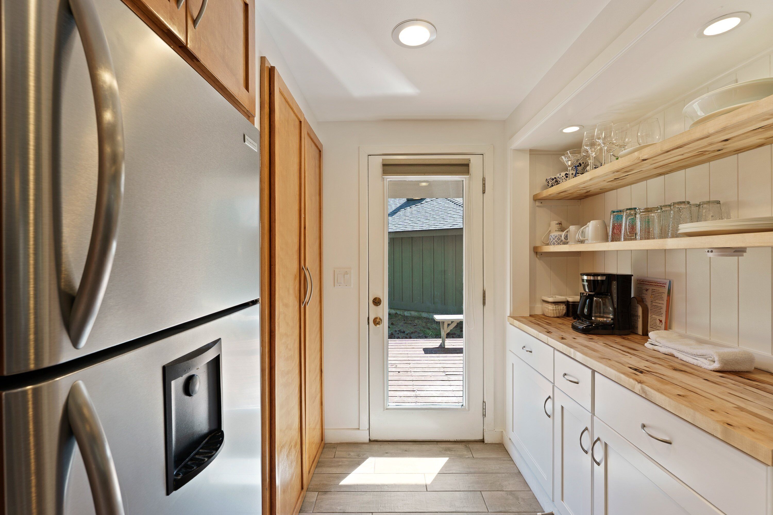 Una cocina con un refrigerador de acero inoxidable, armarios de madera, una encimera blanca y una puerta que da a un espacio exterior.