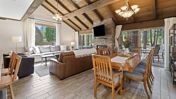 A spacious living area with a high ceiling, wooden beams, a fireplace, a television, and large windows offering a view of the forest.