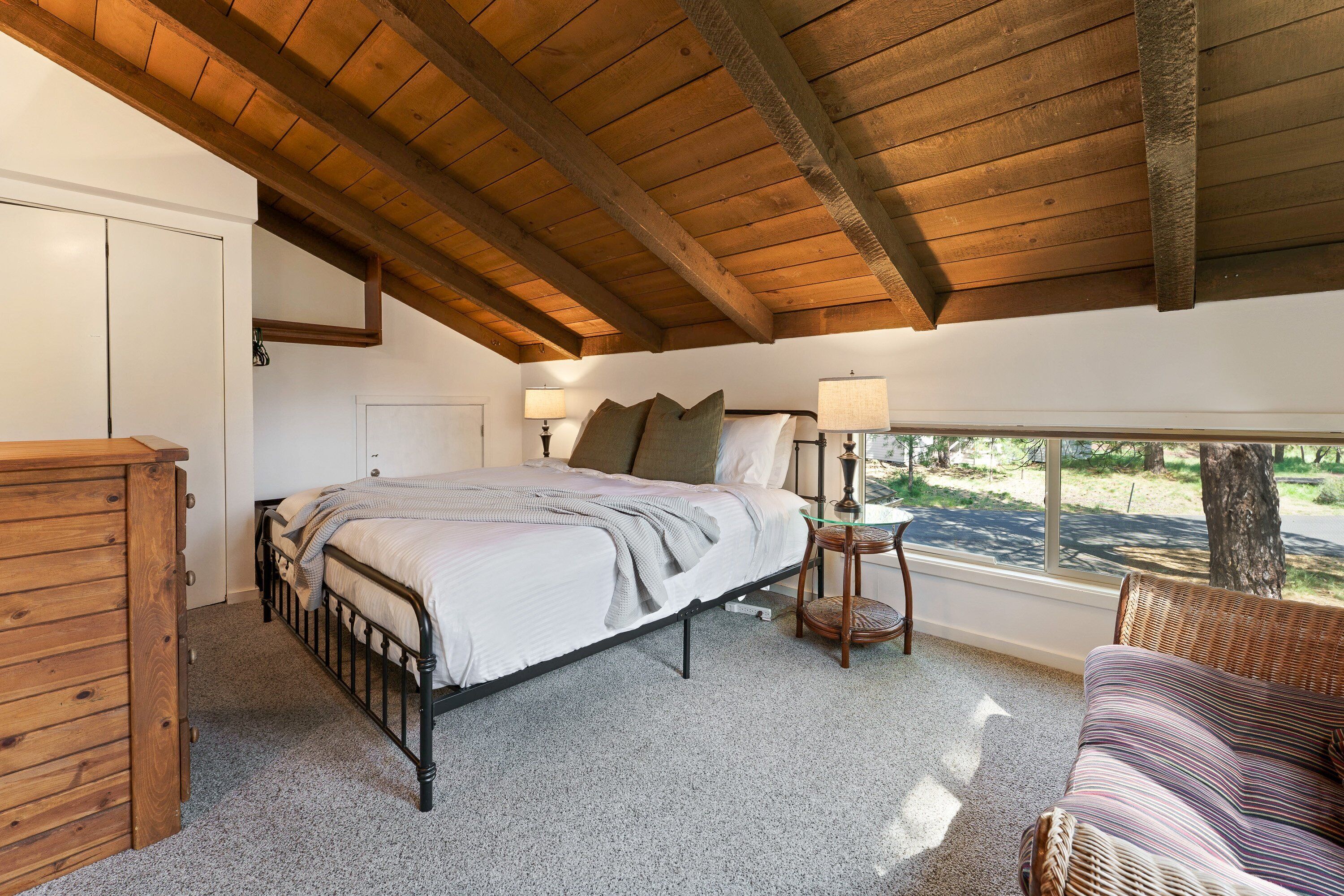 Un dormitorio con cama, cabecera de madera, cómoda, ventana con vistas al exterior y una silla de mimbre.
