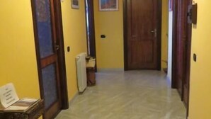 Un couloir aux murs jaunes, doté d’un sol carrelé et de portes en bois. On y trouve un luminaire au plafond, un radiateur et une petite table sur laquelle sont posés des papiers, le tout situé sur le côté gauche.