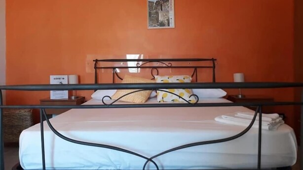 Un llit individual amb estructura metàl·lica, roba de llit blanca i un coixí amb dibuixos grocs. Hi ha una petita taula de nit amb una làmpada i una caixa etiquetada "COVID-19".