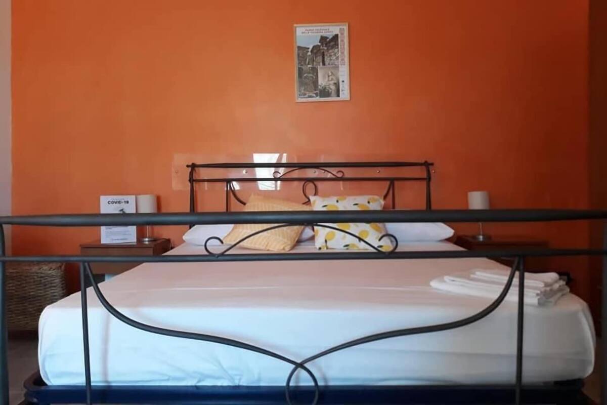 Un lit simple avec un cadre en métal, une literie blanche et un oreiller à motifs jaunes. Il y a une petite table de chevet avec une lampe et une boîte étiquetée « COVID-19 ».