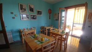 Un espace repas avec des tables et des chaises en bois, un mur bleu orné de tableaux encadrés et une porte-fenêtre coulissante donnant sur un espace extérieur.