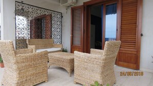 Mobilier en osier comprenant un banc et deux chaises, une porte décorative en métal et des volets en bois sur la façade d’un bâtiment.