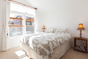 Quarto com uma cama grande, duas luminárias de cabeceira, uma janela com persianas e uma pequena mesa com abajur.
