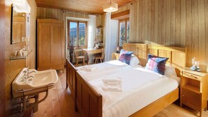 A wooden cabin bedroom with a bed, bedside tables, a mirror, a window, and a bathroom with a sink and mirror. - Hotel du Glacier (Fiesch)
