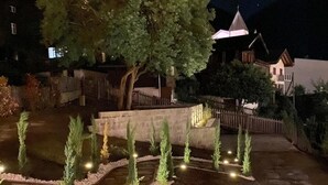 A landscaped garden with a large tree, a stone wall, and a pathway lit by ground lights. - Hotel du Glacier (Fiesch)