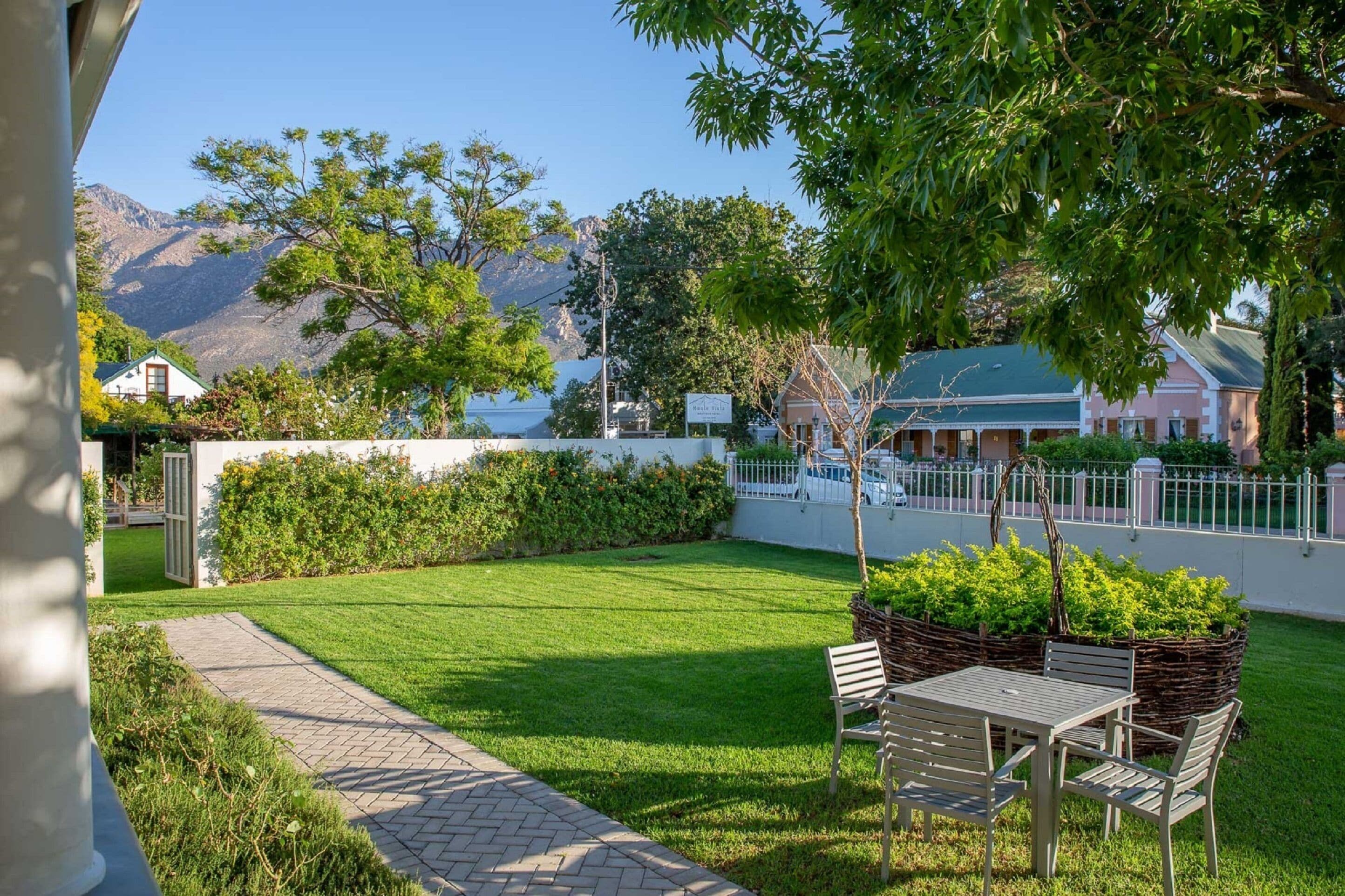 A well-maintained garden with a paved pathway leading to a table and chairs, surrounded by lush greenery and a view of mountains in the background.