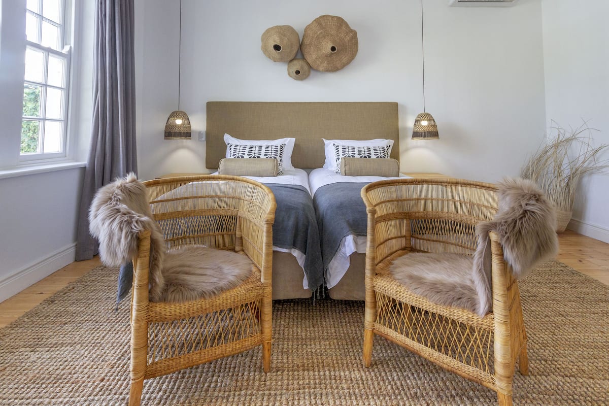Un dormitorio con una cama grande, dos lámparas de noche, un adorno en la pared y dos sillones de mimbre con mantas de piel.