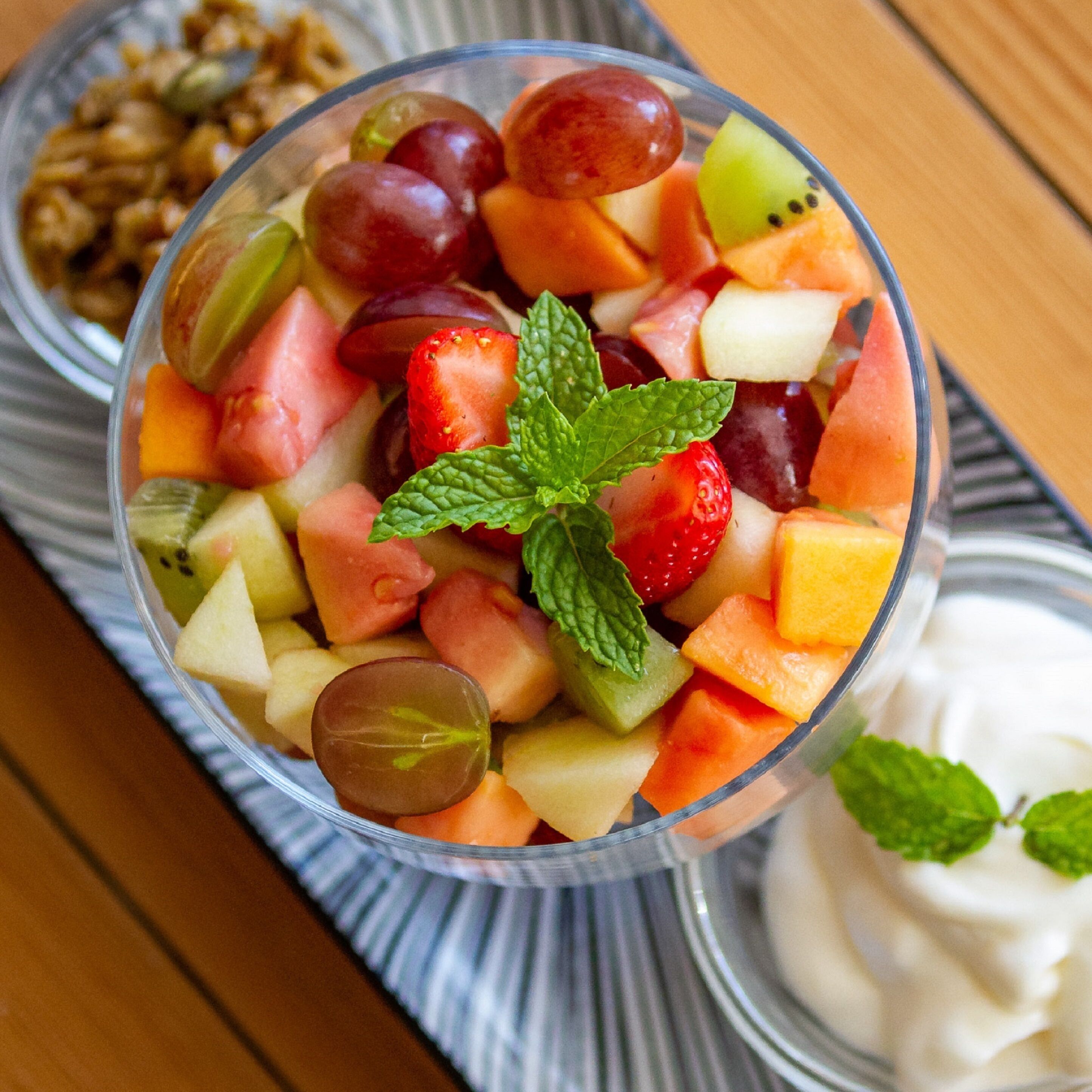 A fruit salad in a clear glass bowl with a mint garnish, accompanied by a side of yogurt and granola.