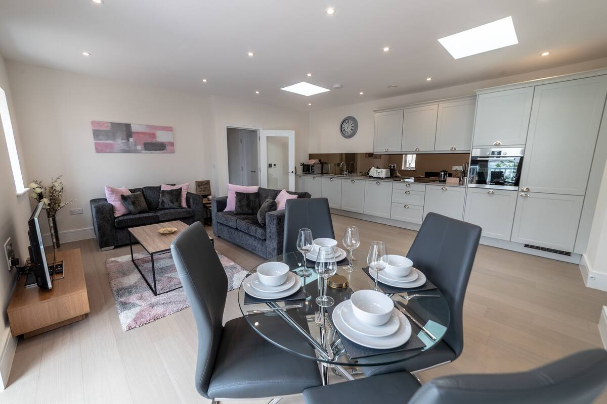 A modern living room with a dining table set for four, a grey sofa, and a kitchen area with white cabinets and a clock.