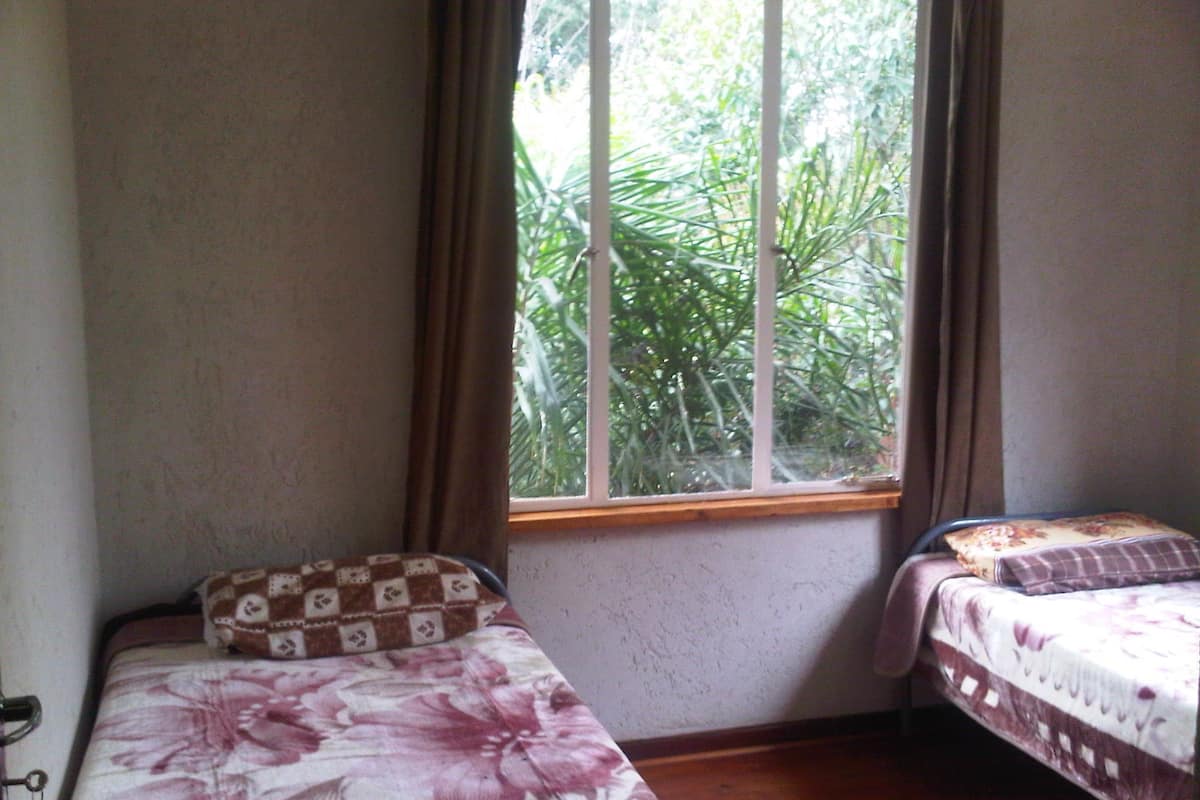A small room with two beds, one with a floral bedspread and the other with a patterned bedspread. There is a window with curtains, and the room has a wooden floor.