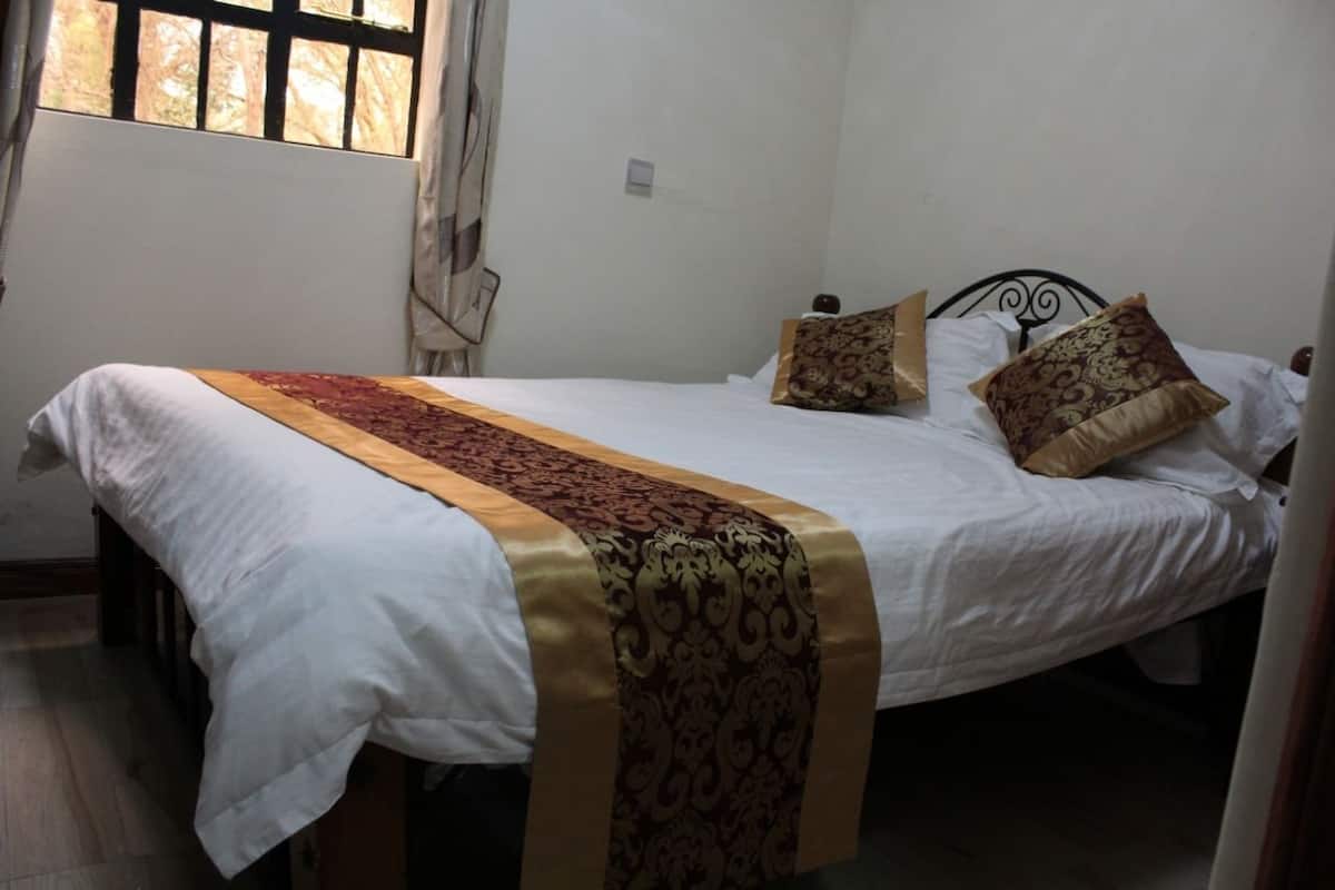 A single bed with a white and gold bedspread, two patterned pillows, and a window with curtains.