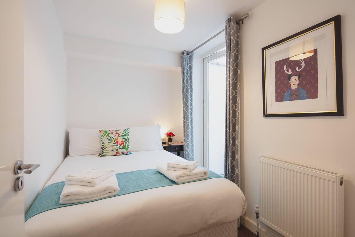 A neatly made bed with white linens and a teal throw, a framed picture on the wall, and a window with curtains.