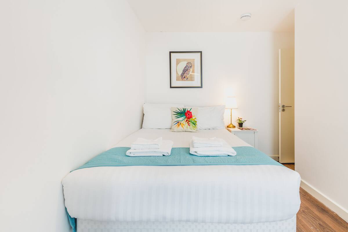 A neatly made bed with white linens and a teal throw blanket, a framed artwork on the wall, and a bedside table with a lamp and a small plant.