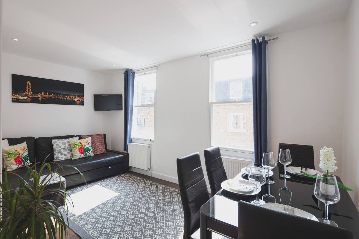 A modern living room with a black sofa, a dining table set for two, and a large window with curtains.
