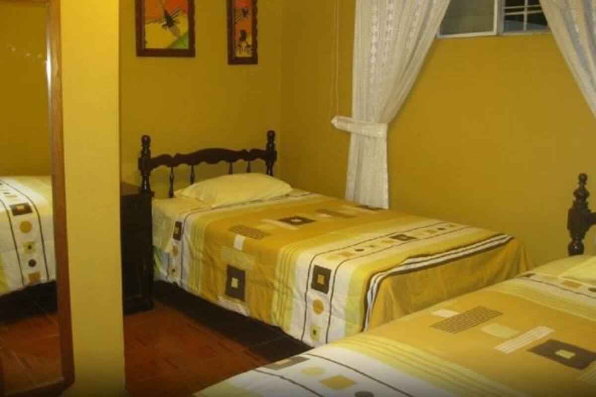 Une chambre avec deux lits, une tête de lit en bois, une fenêtre avec des rideaux blancs et deux tableaux encadrés accrochés au mur.