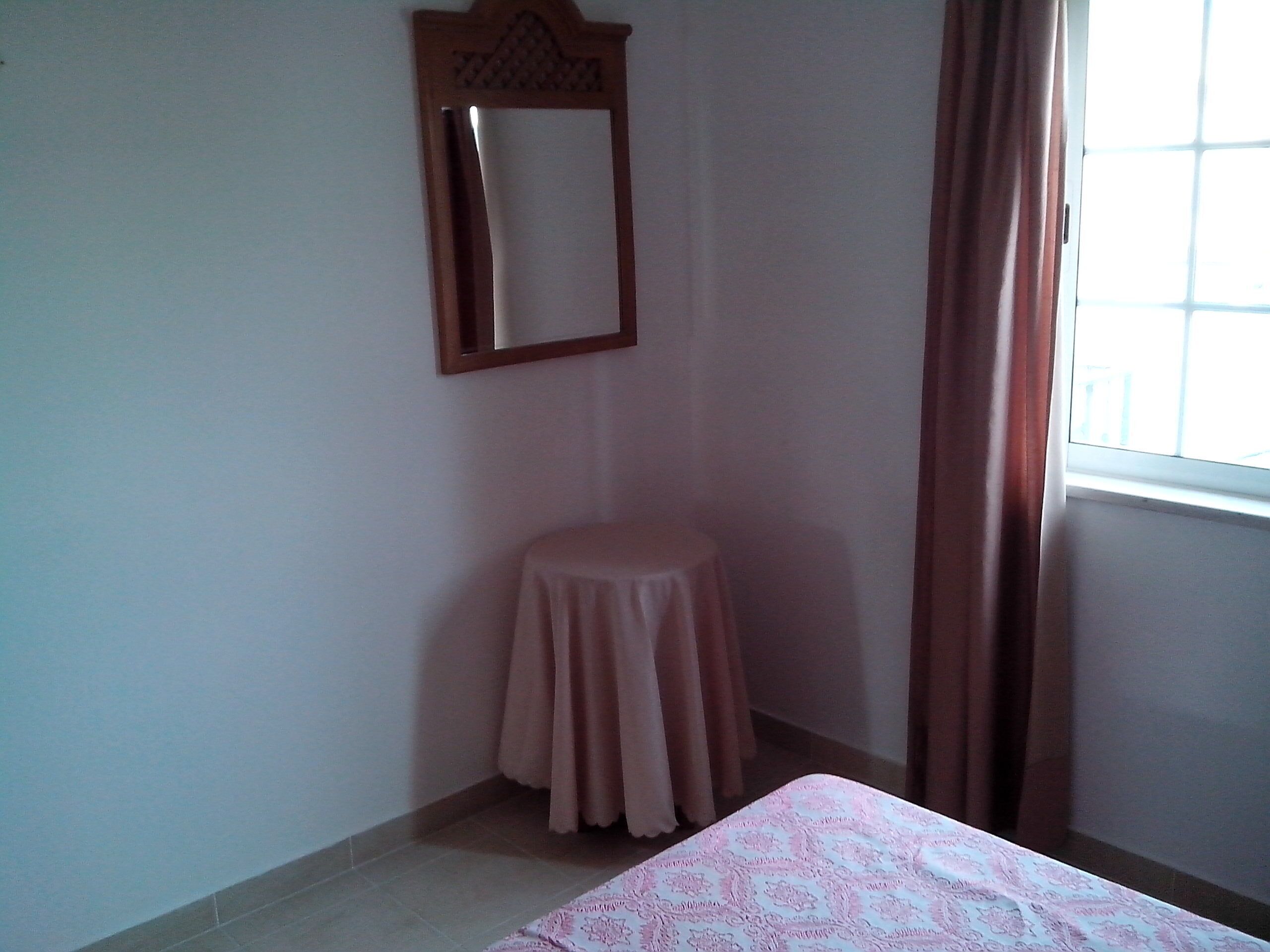 A small room with a bed, a stool, a mirror, and a window with curtains.