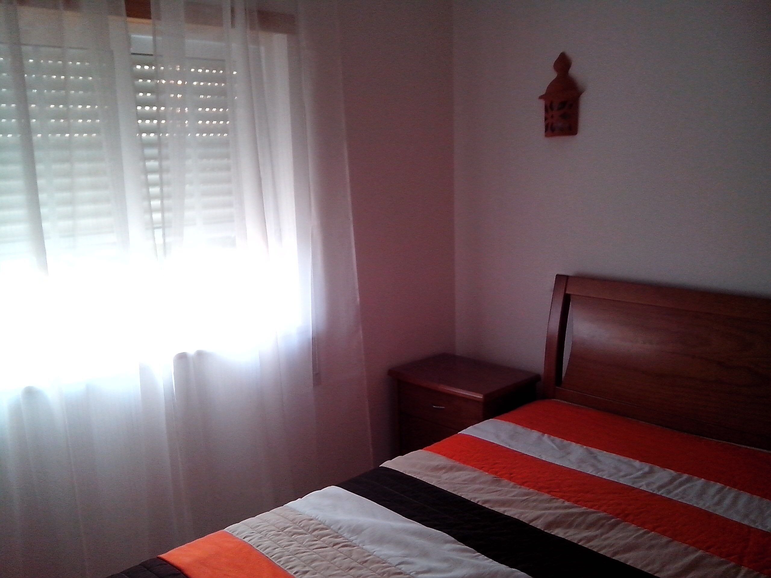 Ein Schlafzimmer mit Bett, Nachttisch, Fenster mit transparenten Vorhängen und einer Wandleuchte.