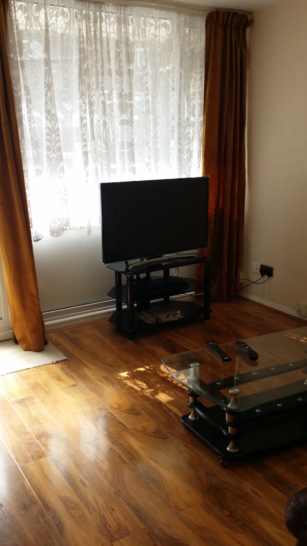 Uma sala de estar com uma televisão de tela plana sobre um suporte preto, uma mesa de centro de vidro e piso de madeira.