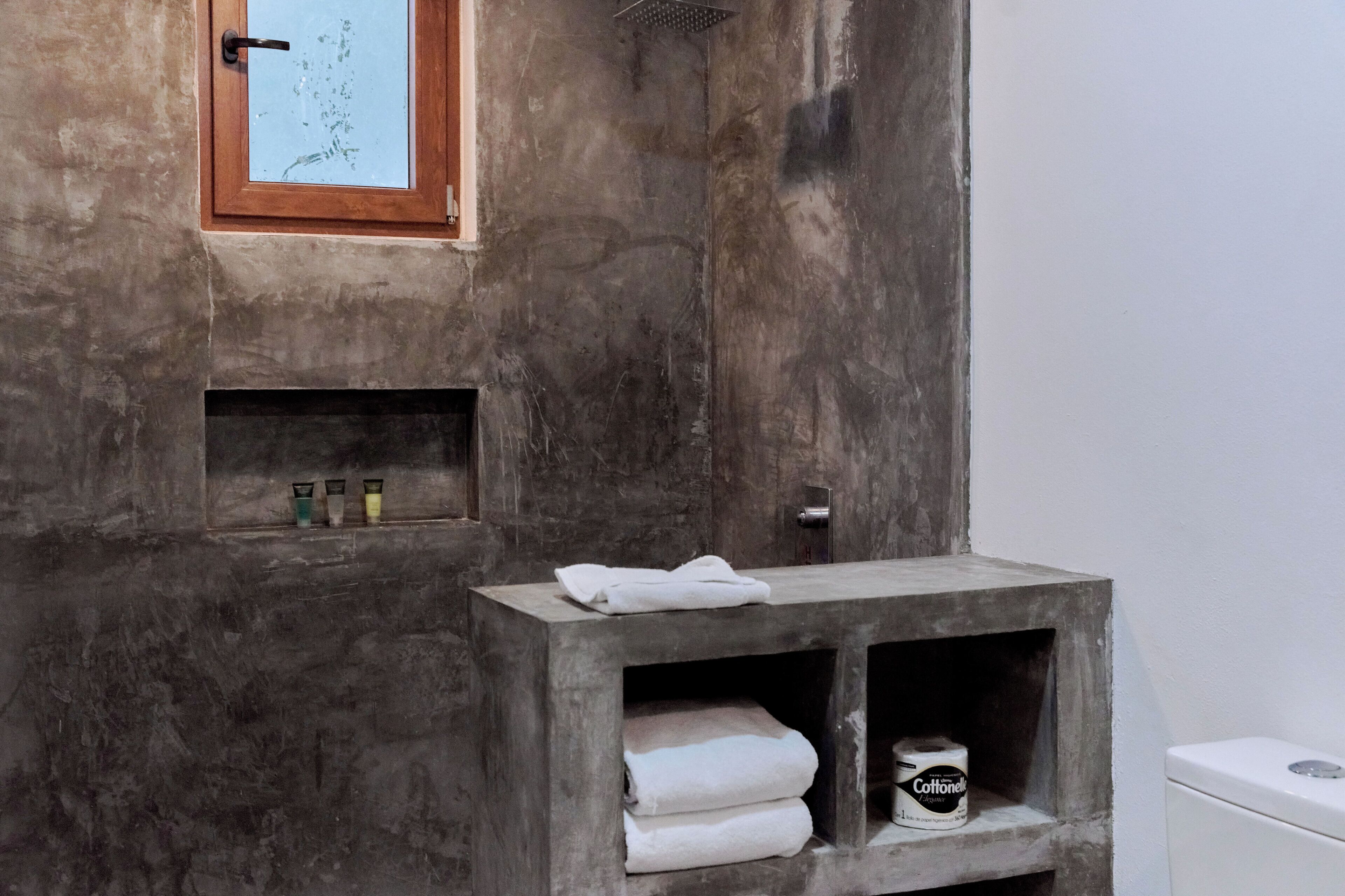 A bathroom with a concrete wall, a window, a sink with a towel, and a toilet.