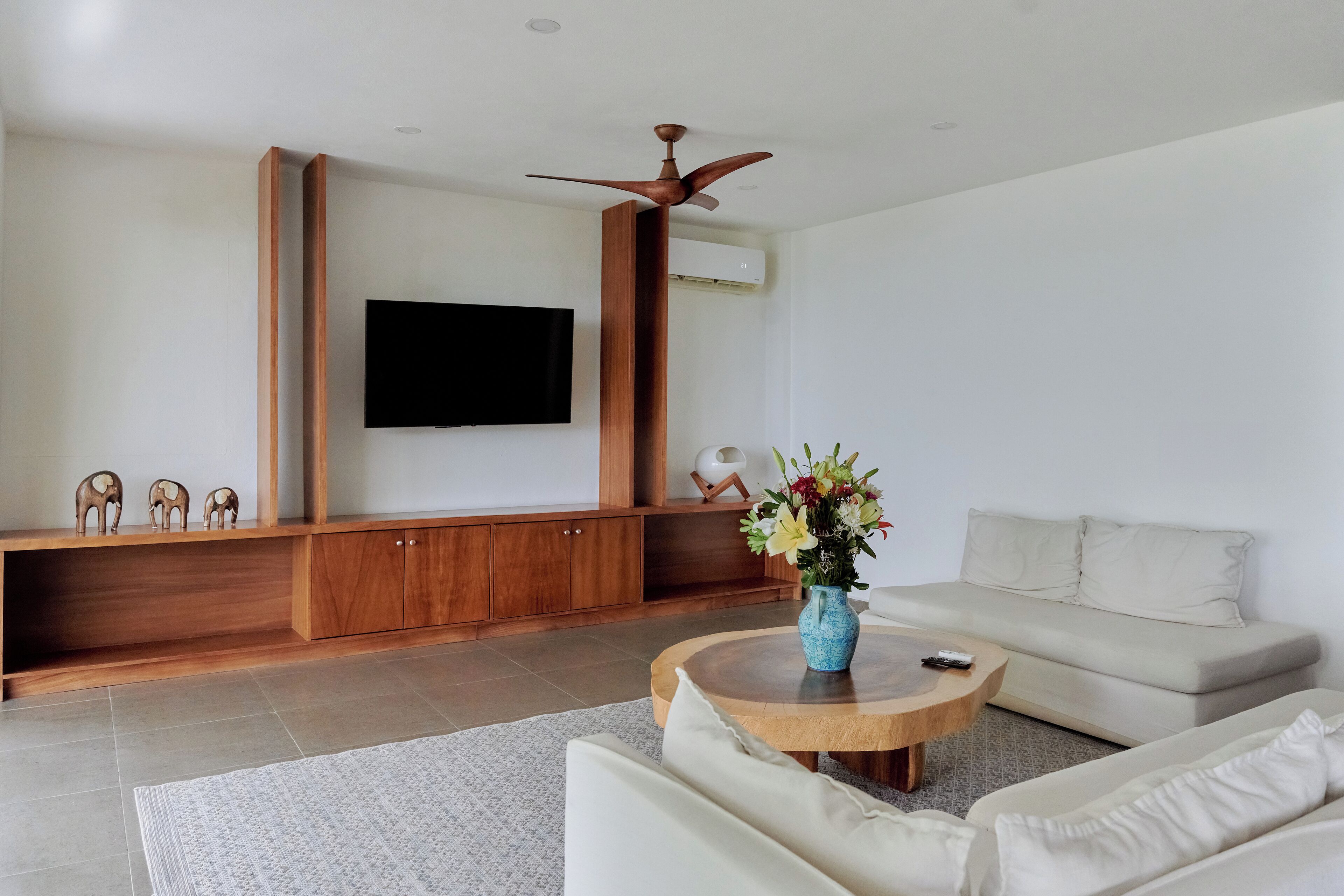 Een moderne woonkamer met een flatscreen televisie aan de muur, een houten tv-meubel, een beige bank en een ronde salontafel met een bloevas.