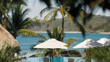 Zona junto a la piscina con sillones de descanso y sombrillas, con vista a un cuerpo de agua y palmeras.