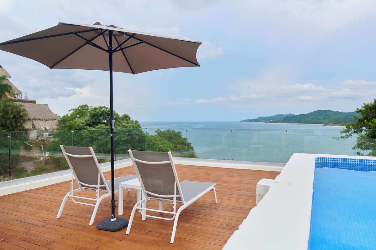 Terraza de madera con dos sillones de descanso y una sombrilla grande, con vista a una piscina y un paisaje de la costa y colinas.