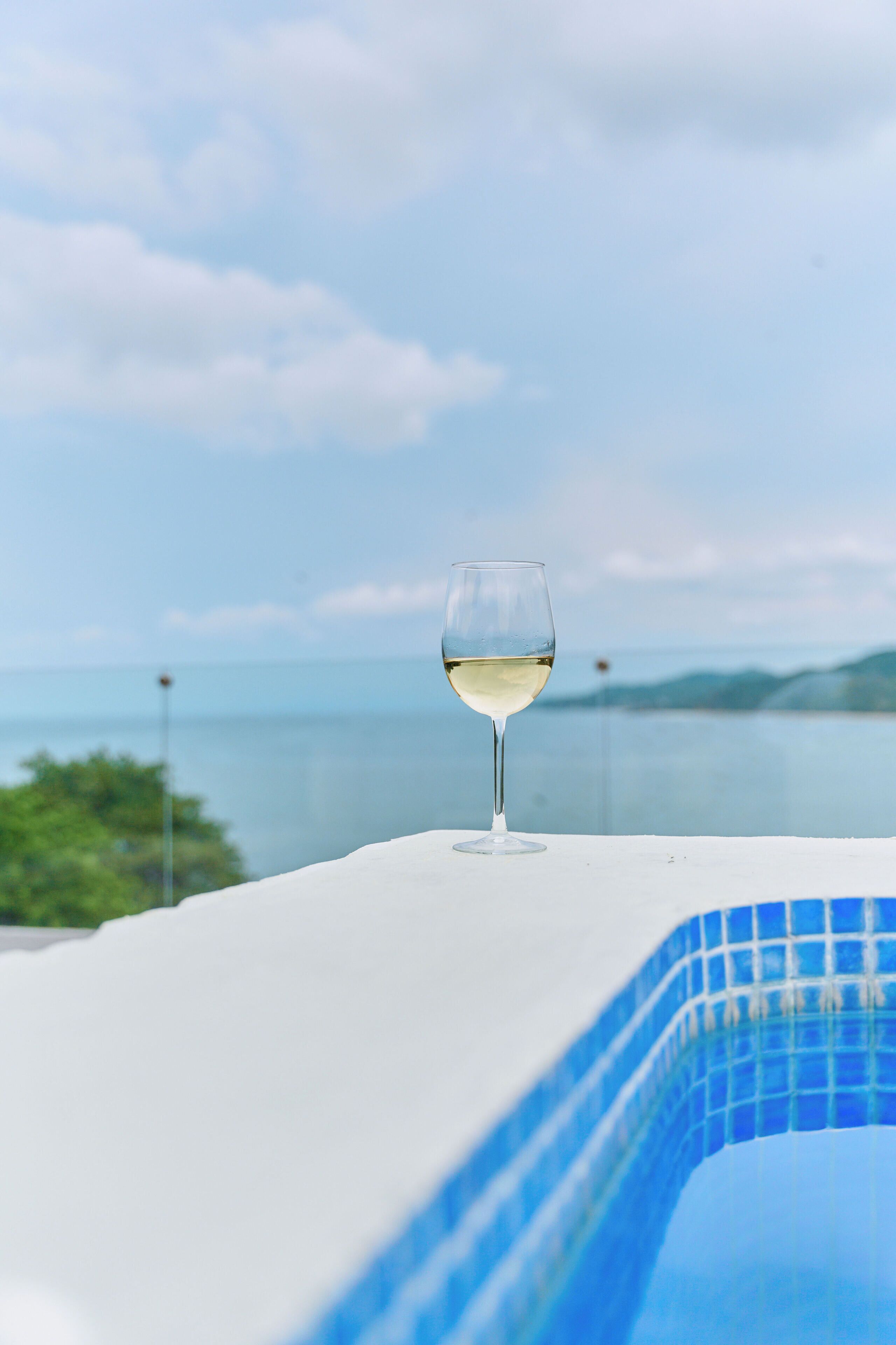 Um copo de vinho branco sobre superfície branca, com piscina e vista para o mar e colinas ao fundo.