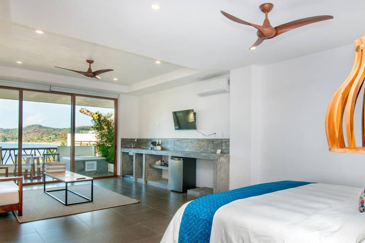 Habitación de hotel moderna con una cama grande, zona de comedor con vistas, cocina y dos ventiladores de techo.