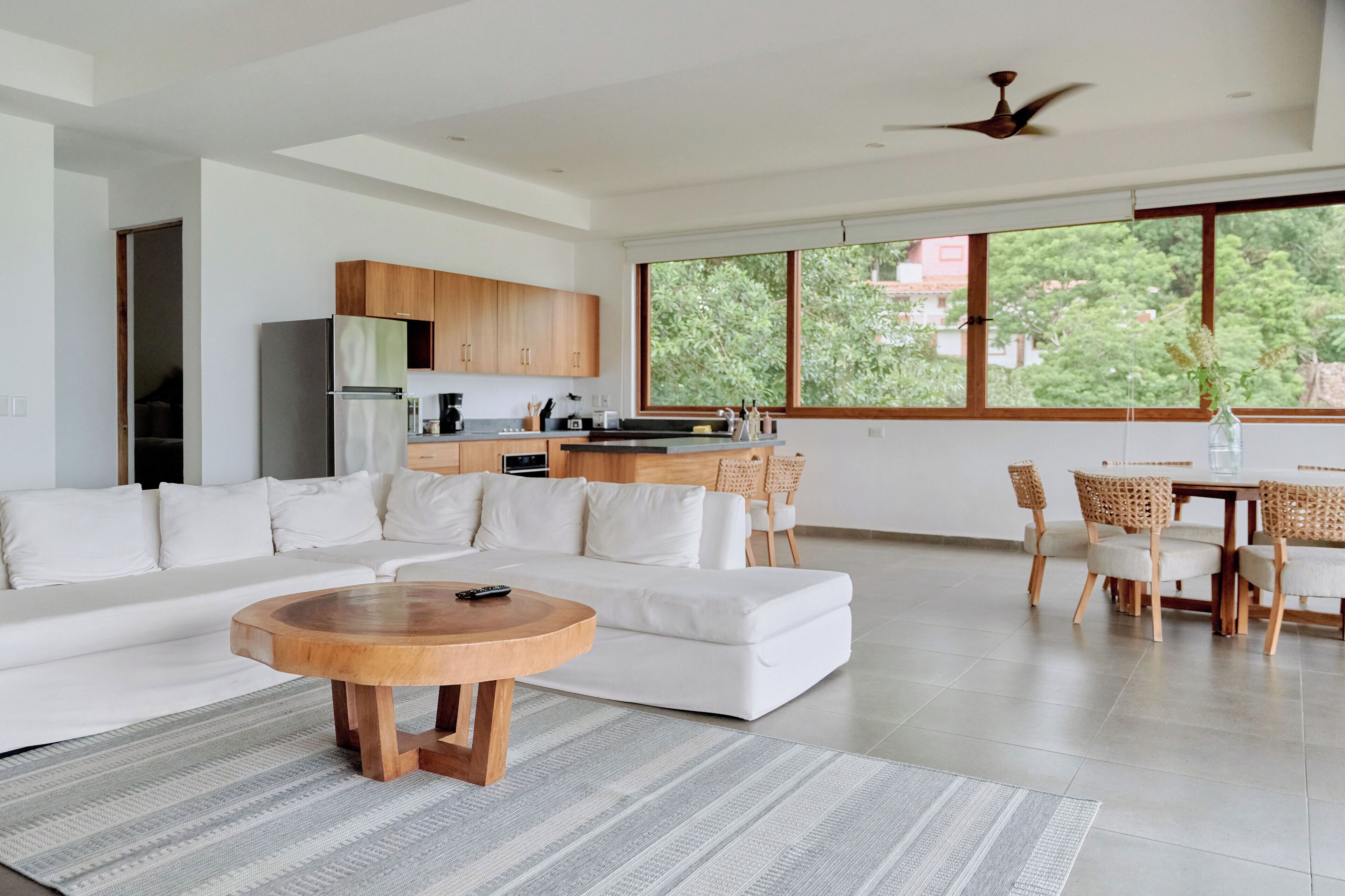 Un espacio moderno para vivir con un sofá seccional blanco, una mesa de centro de madera, una cocina con armarios de madera, un comedor con sillas de mimbre y grandes ventanales con vistas a la vegetación.