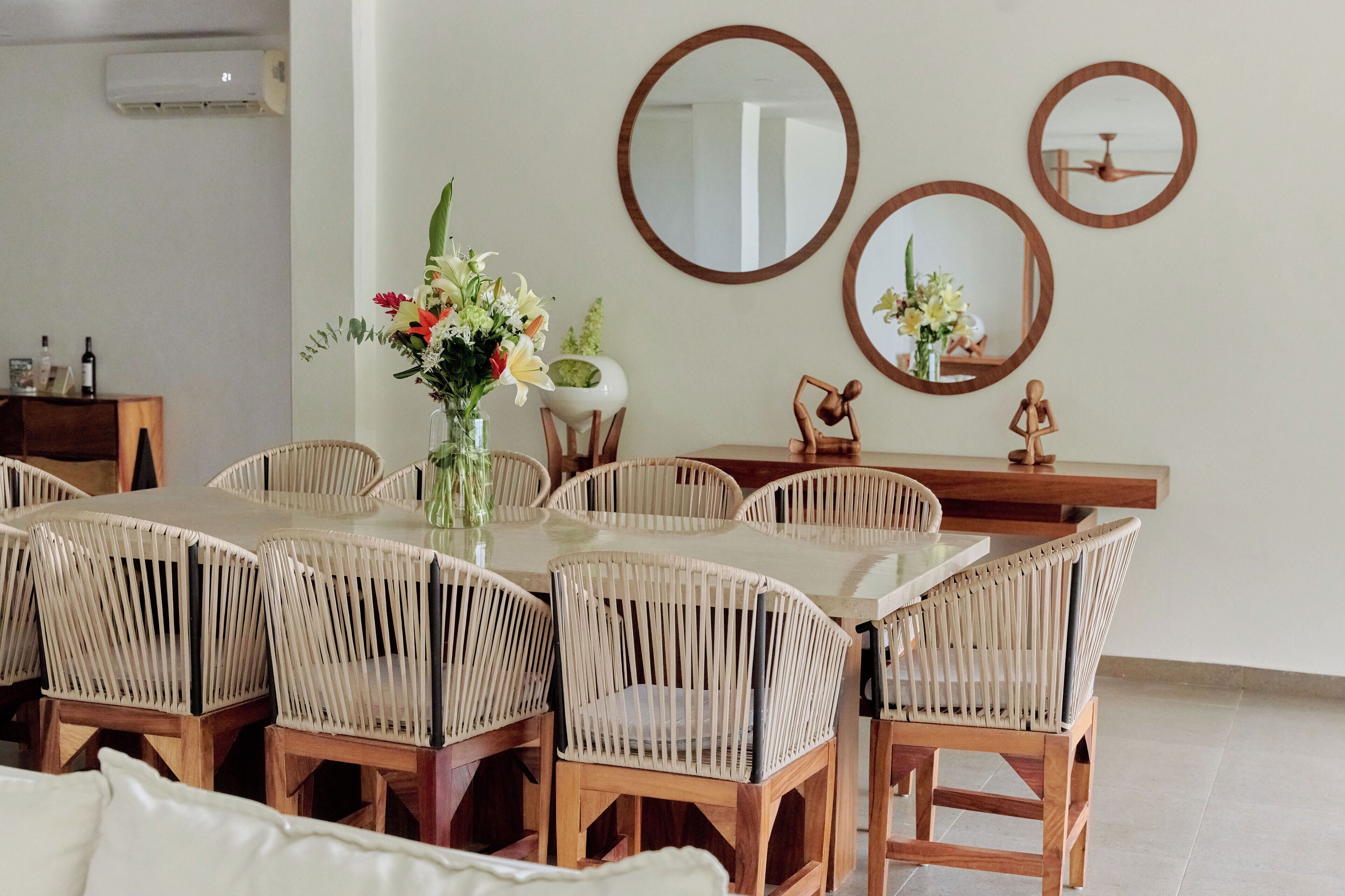 A dining area with a round table and wicker chairs, a vase with flowers, and three wall-mounted mirrors.