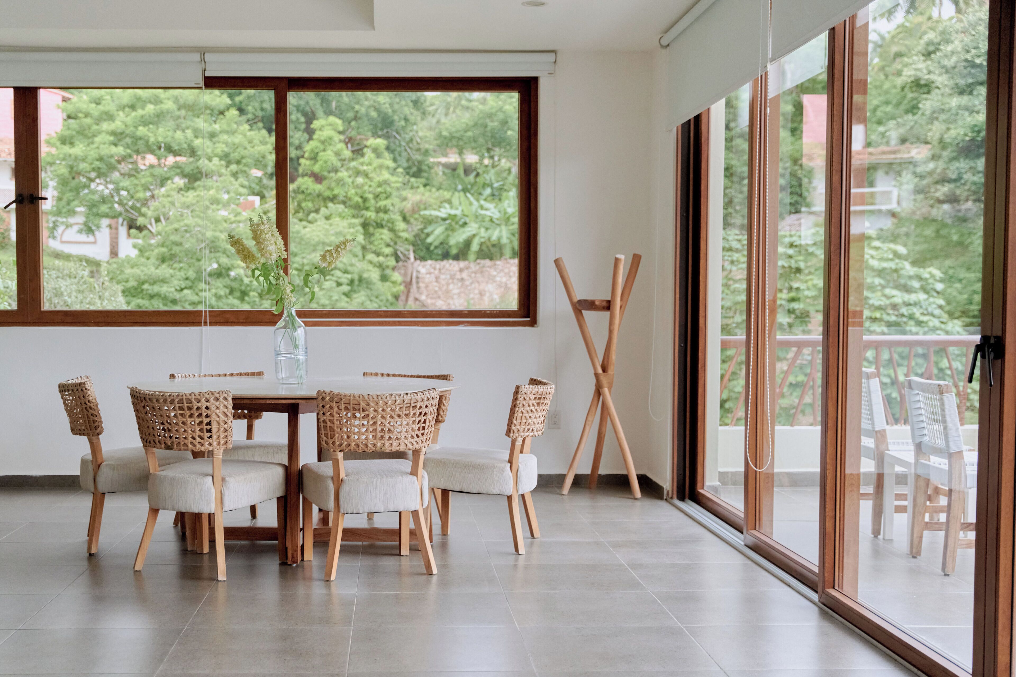 A dining area with a table and chairs, a vase with flowers, and a view of greenery through large windows.